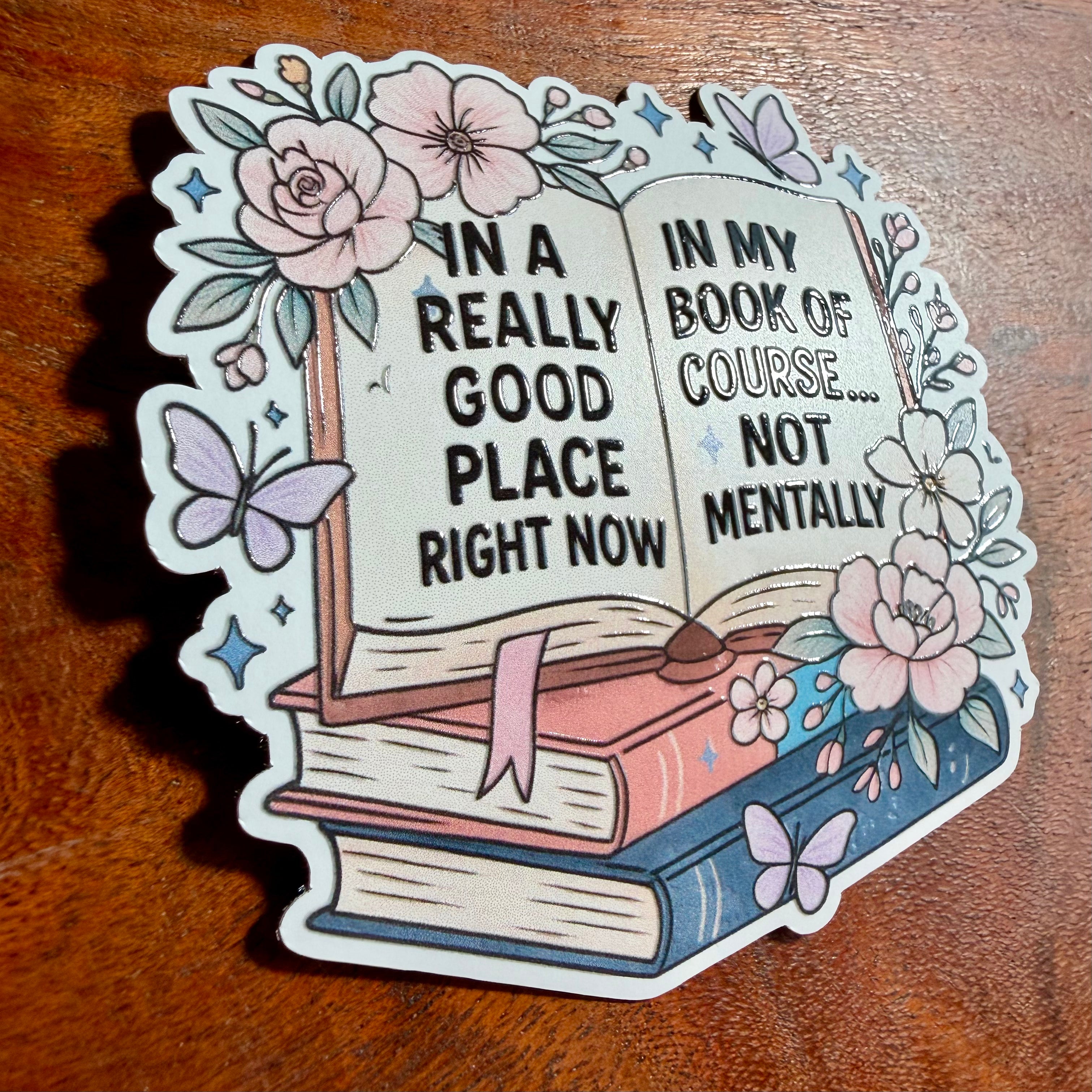 Decorative sticker with an open book design, flowers, and butterflies on a wooden surface.
