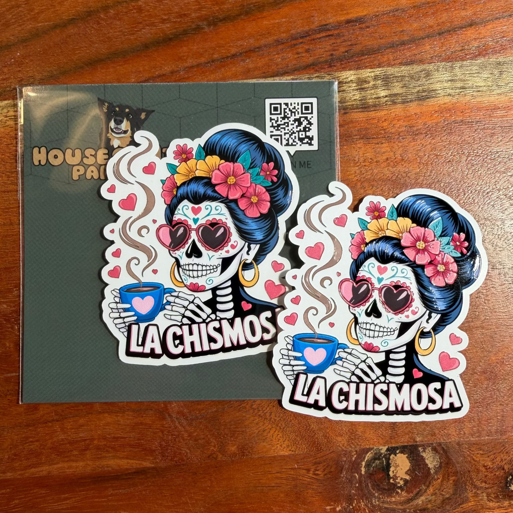 Two colorful stickers of a skeleton with flowers on a green card, placed on a wooden surface.