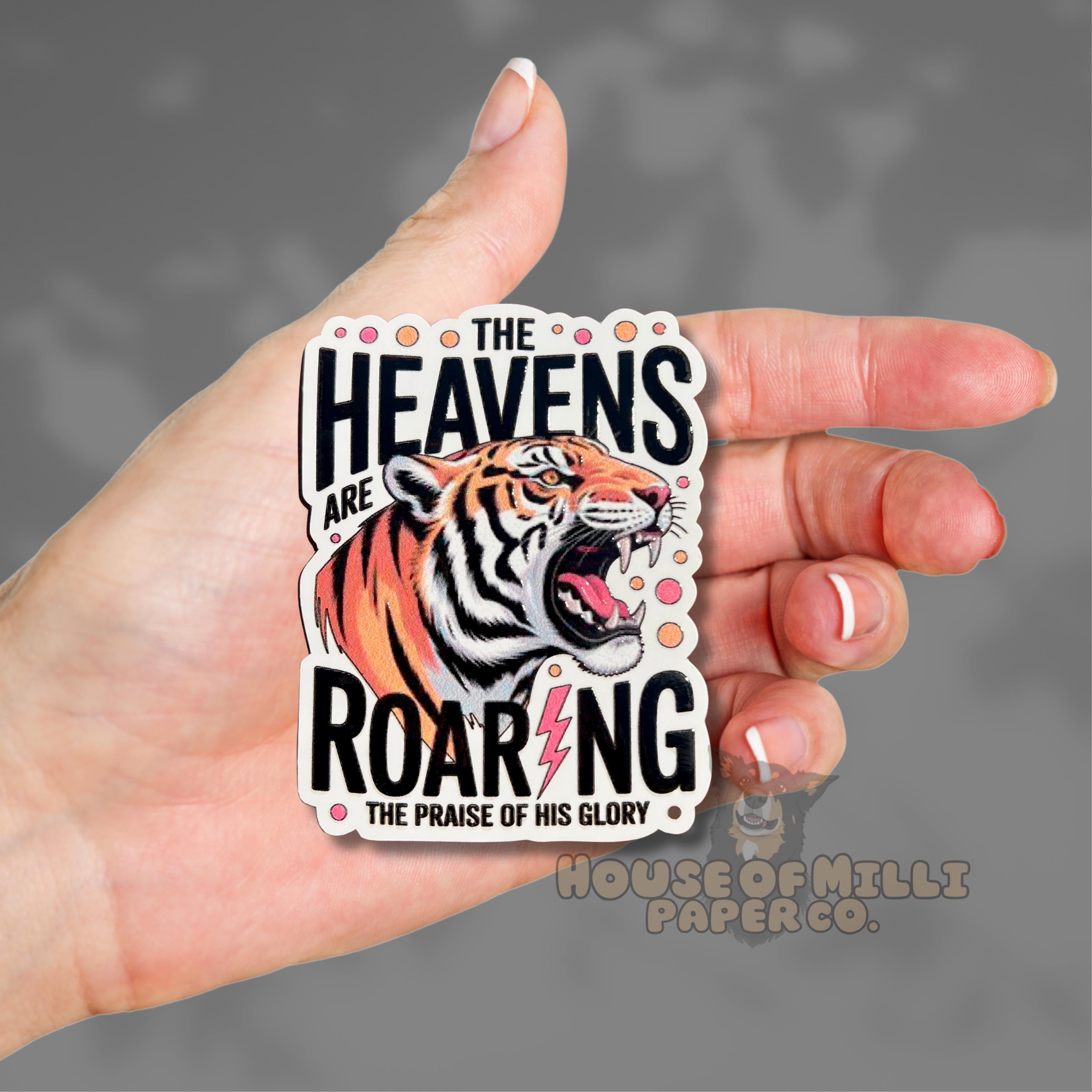 Hand holding a sticker with a tiger design and text 'The Heavens are Roaring' against a gray background.