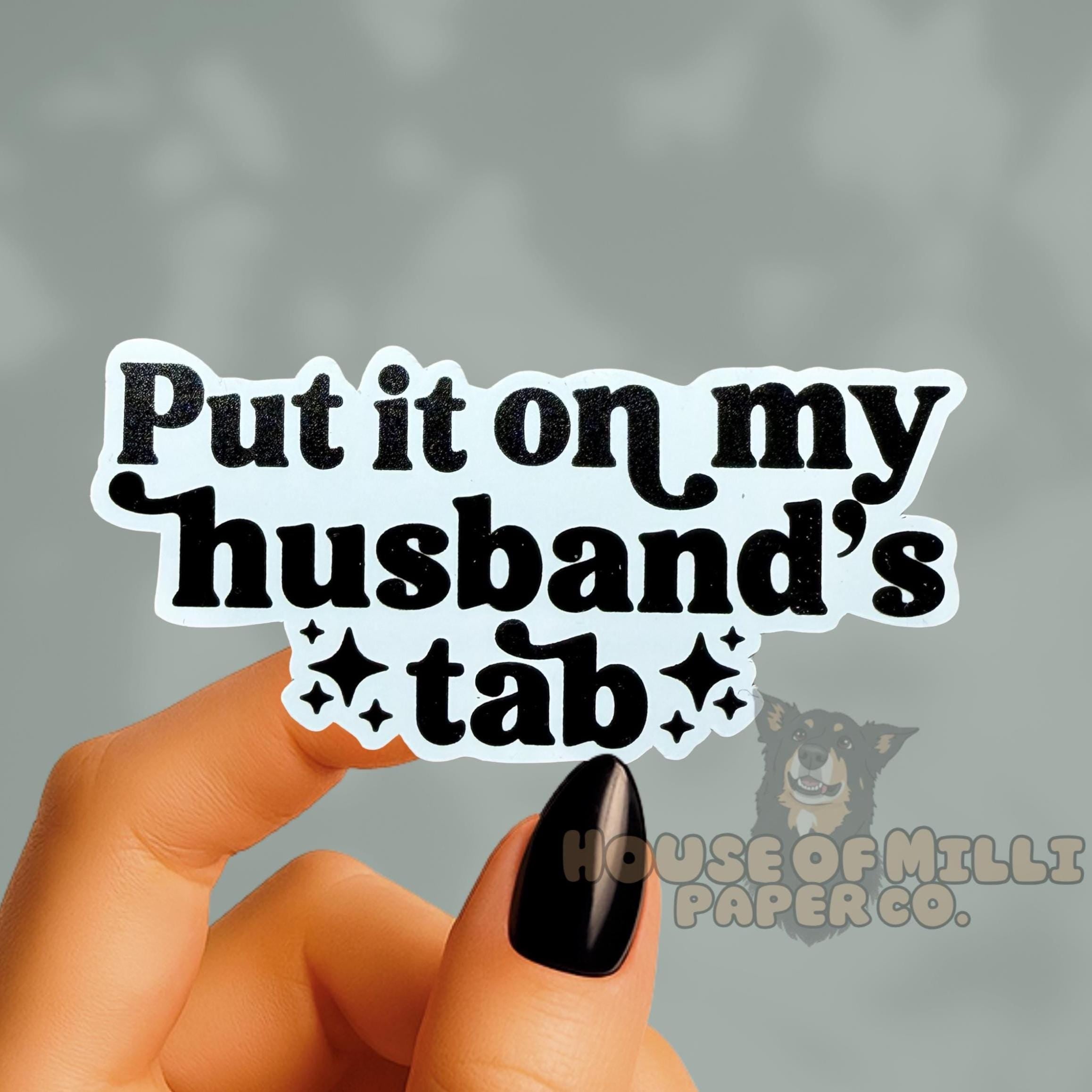 Hand holding a sticker with text 'Put it on my husband's tab' against a gray background.
