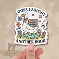 Sticker with a duck holding books and text 'Oops, I bought another book' held by a hand.