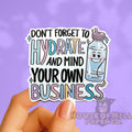 Sticker with text 'Don't forget to hydrate and mind your own business' held by a hand on a purple background.