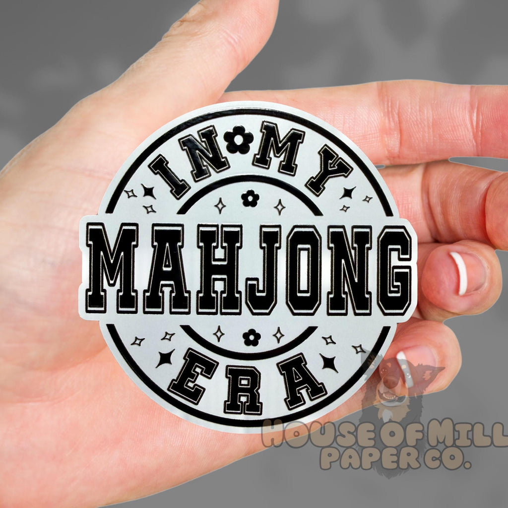 Hand holding a round sticker with 'IN MY MAHJONG ERA' text on a gray background