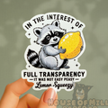 Sticker with a raccoon holding a lemon and text, held by a hand against a green background.