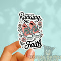 Sticker with pink sneakers and 'Running on Faith' text held by a hand against a light blue background.