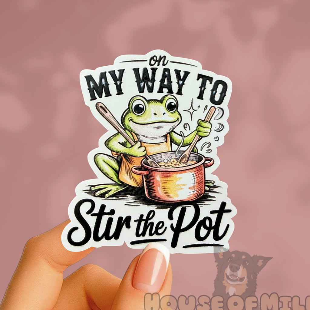 Sticker with a frog design and text held by a hand against a pink background