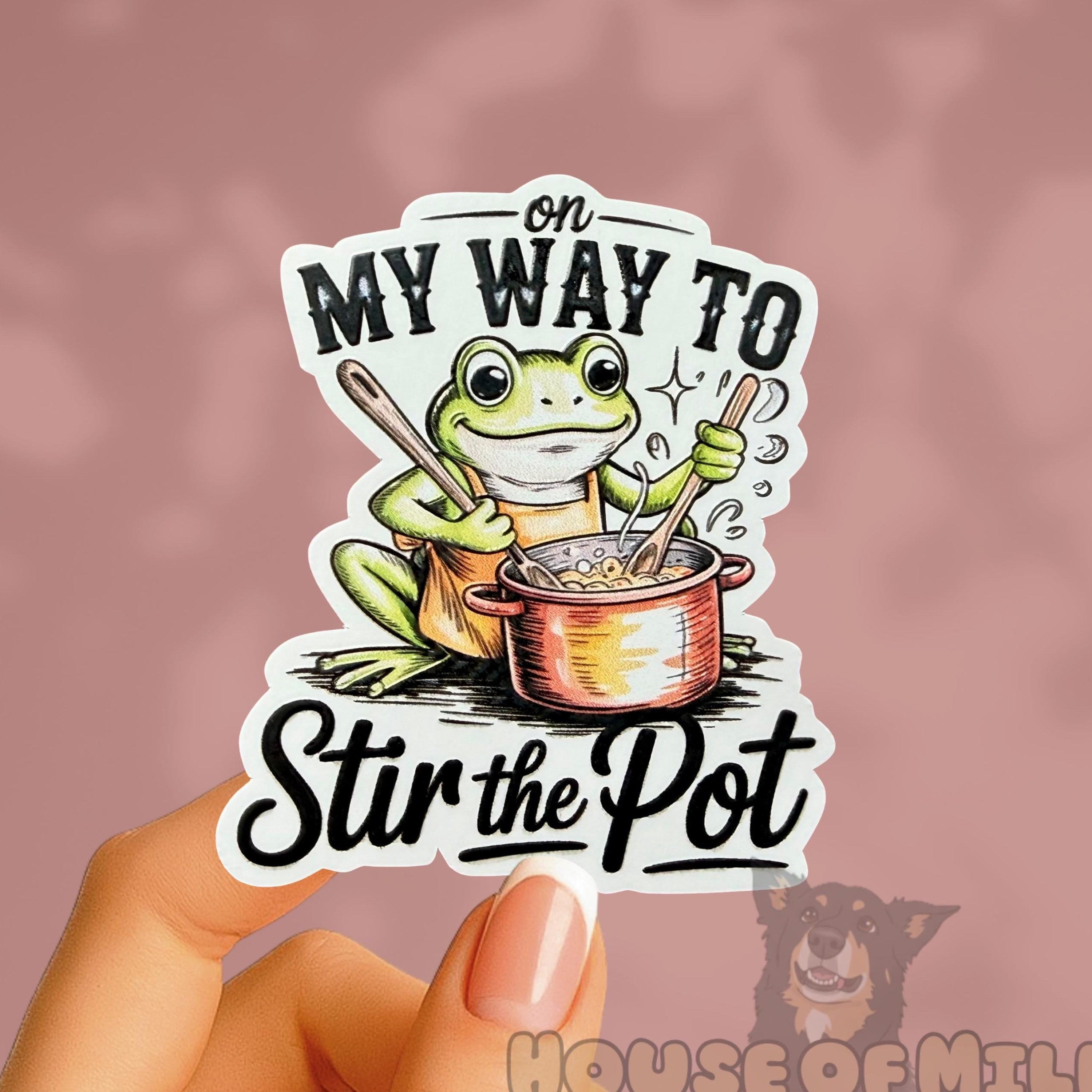Sticker with a frog design and text held by a hand against a pink background