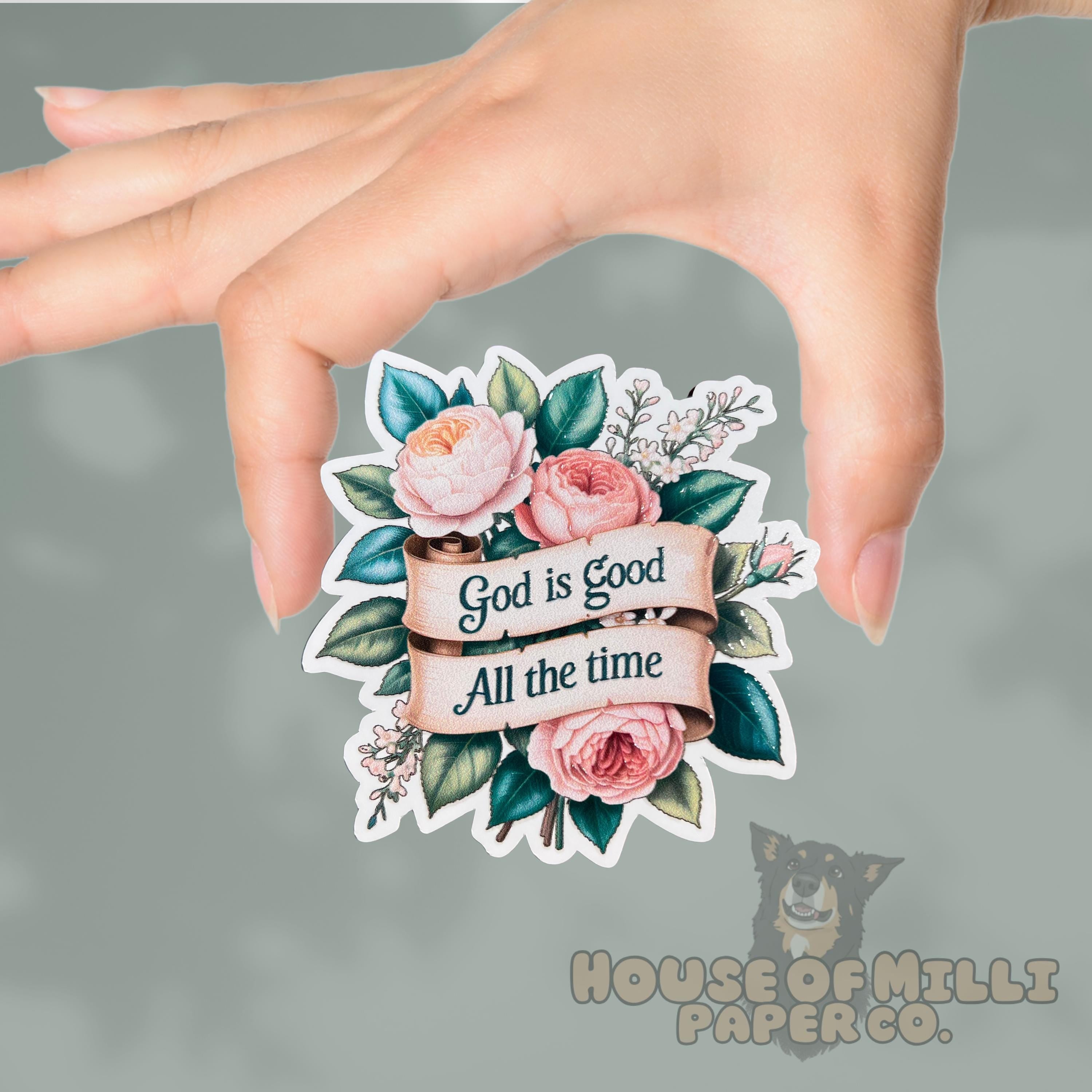 Hand holding a decorative sticker with floral design and text 'God is Good All the time' on a gray background.