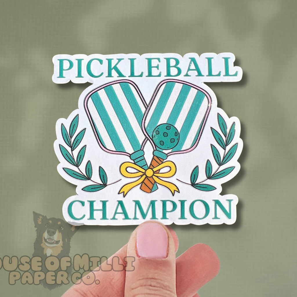Sticker with pickleball design and 'Pickleball Champion' text held by a hand on a green background.