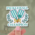 Sticker with pickleball design and 'Pickleball Champion' text held by a hand on a green background.