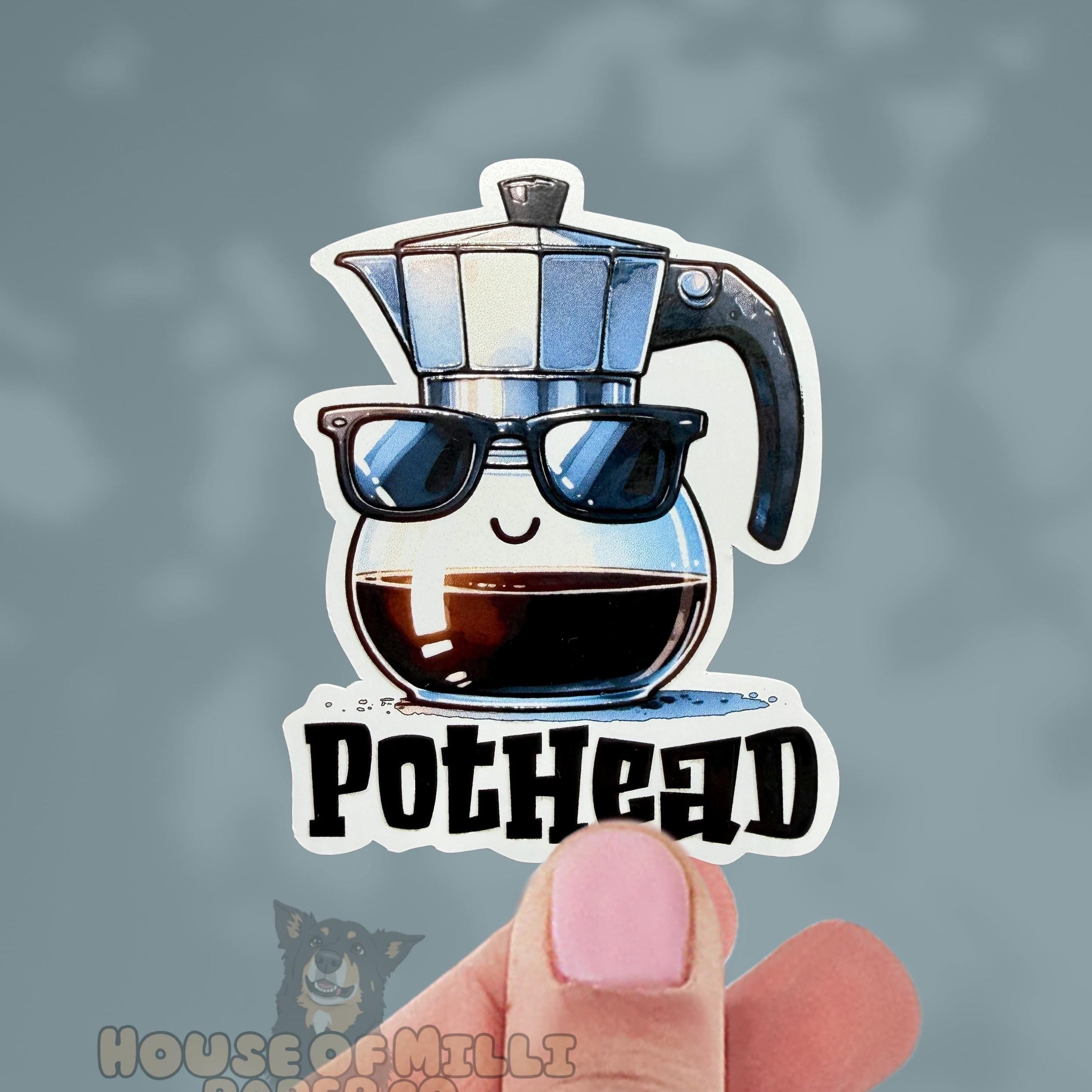 Sticker of a coffee pot with sunglasses and 'POTHEAD' text held by a hand against a blurred background.