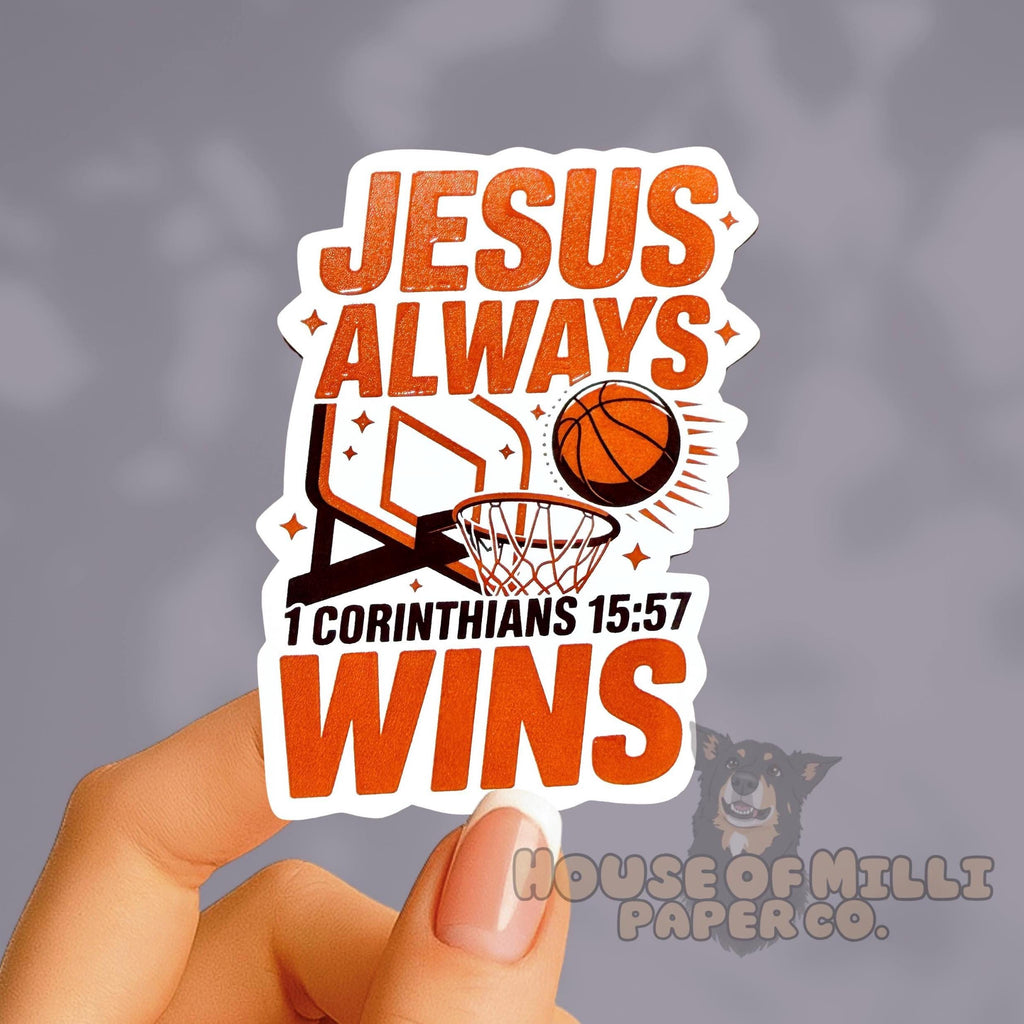 Sticker with 'Jesus Always Wins' text and basketball graphics held by a hand against a gray background.
